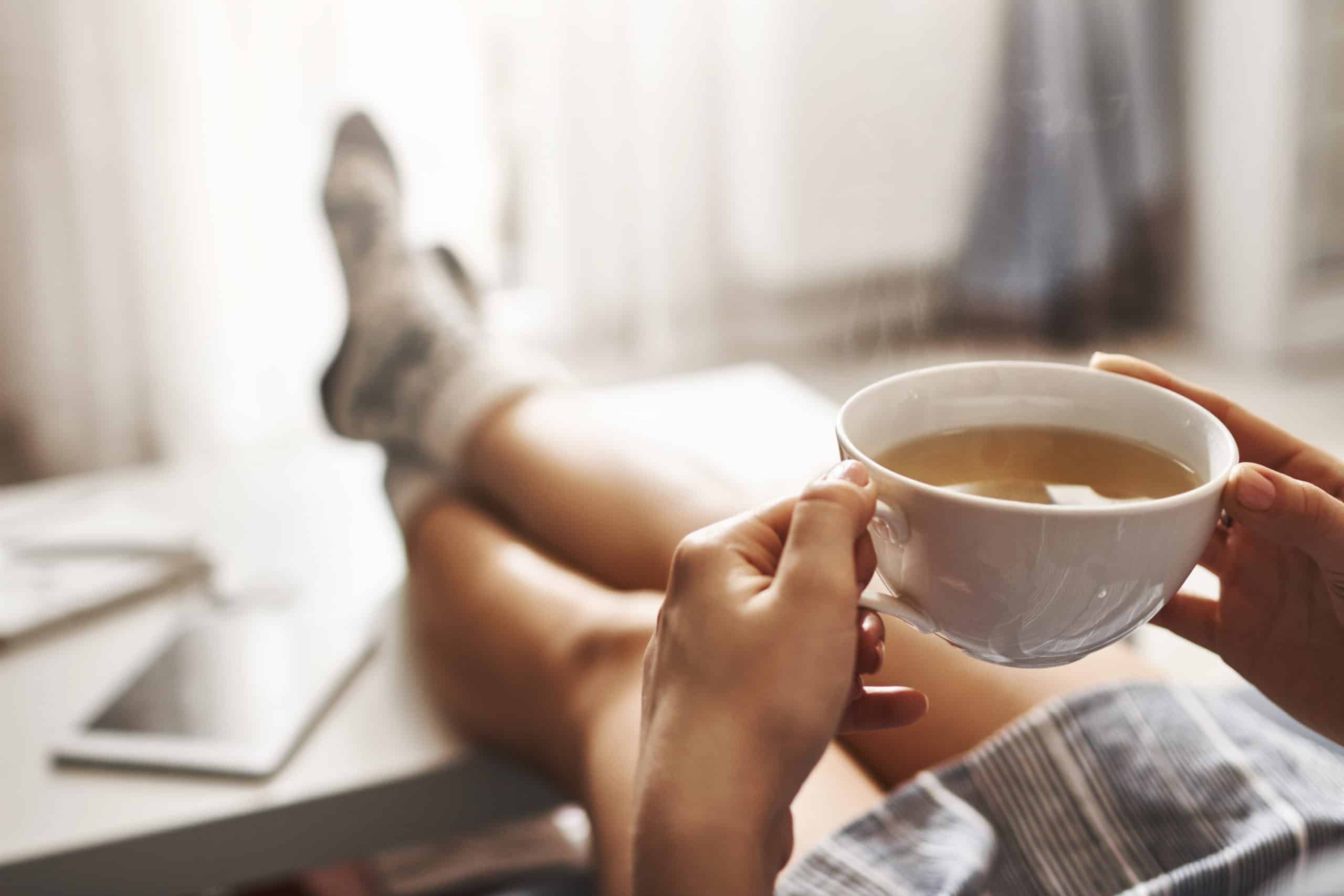 Whitney Sleep blog banner features woman in oversized shirt takes break at home holds a cup of tea while lying on couch, holding legs on coffee table, drinking hot coffee and enjoying morning, being in dreamy and relaxed mood.