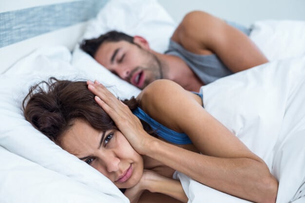 Whitney Sleep blog banner features woman awake in bed while man snores. She blocks her ears with hands as her partner slumbers.