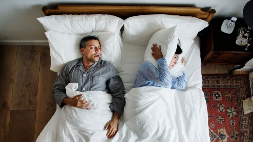 A man being treated for sleep apnea at Whitney Sleep in Minnesota sleeps with his mouth open while the woman next to him covers her ears with her pillow.