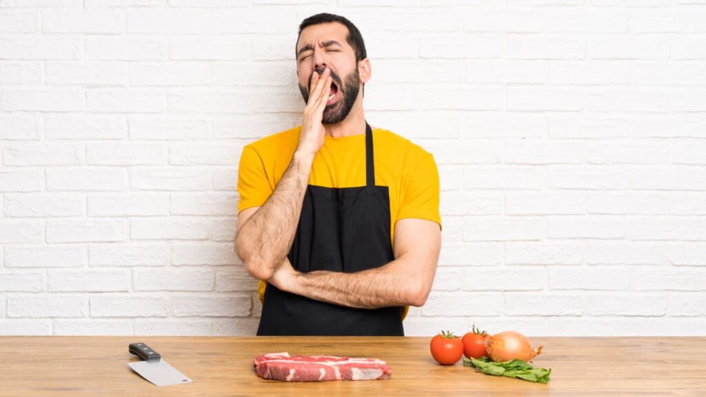 A man standing behind a cutting board and a piece of meat yawns.