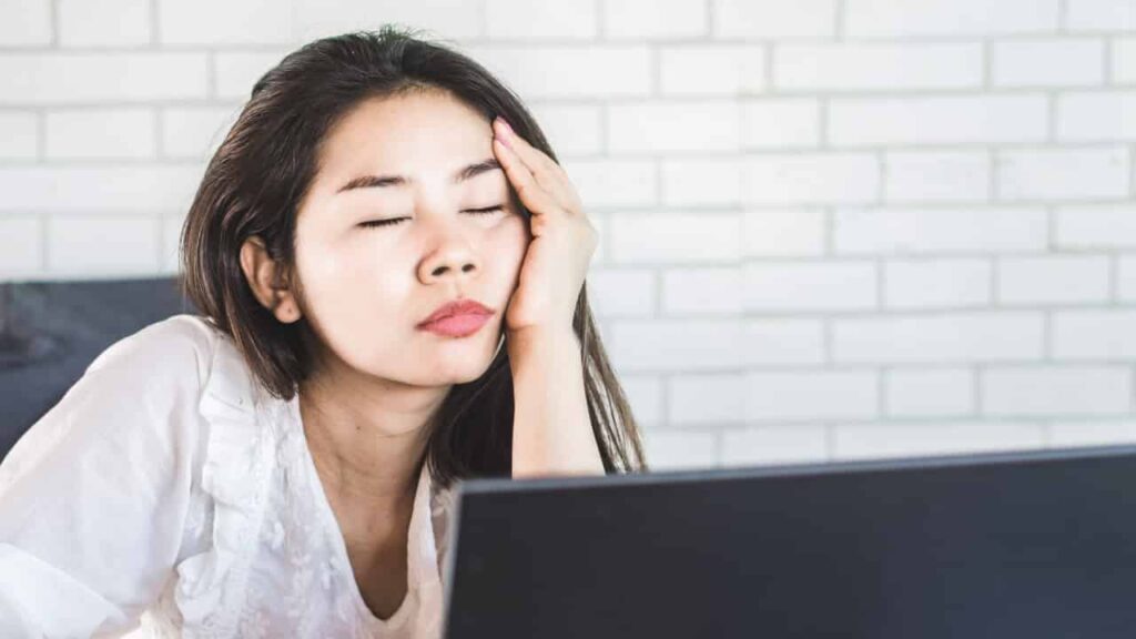 A young woman appears to fall asleep at her computer. Her eyes are closed and her head rests in her hand.