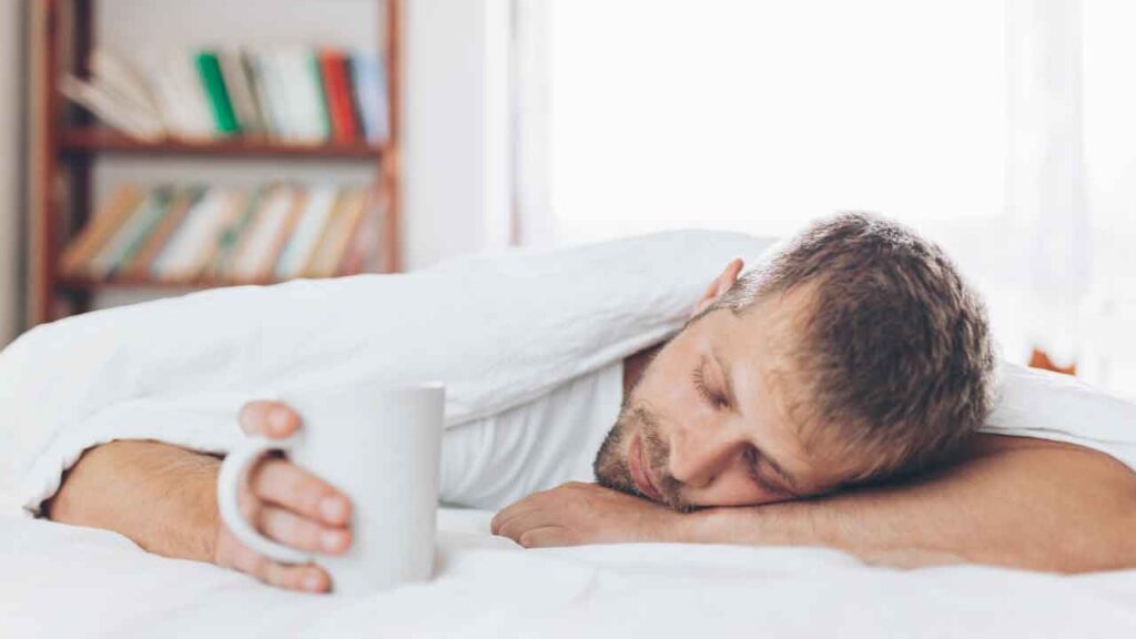 Photo of a man who appears to have fallen asleep holding a cup of coffee. He lies on a bed with head resting on his arm like a pillow.