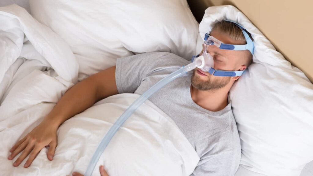 A young man sleeps in a comfy bed. He wears a CPAP machine.