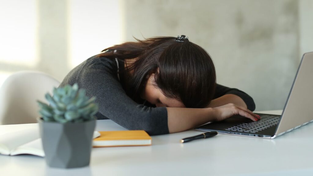A woman has fallen asleep at a table, her head resting on one arm, her other arm still on top of a laptop keyboard.