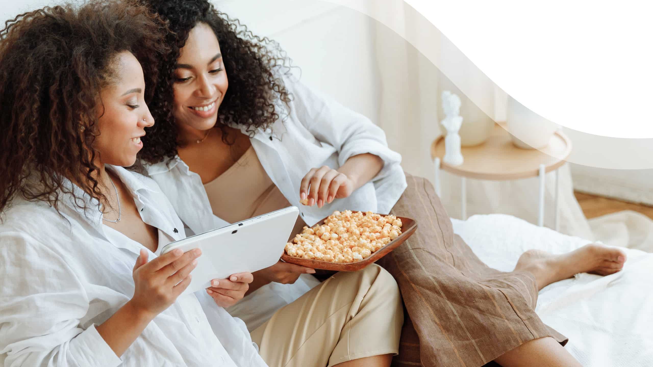 A pair of women on a couch eat popcorn together as they watch something on a tablet