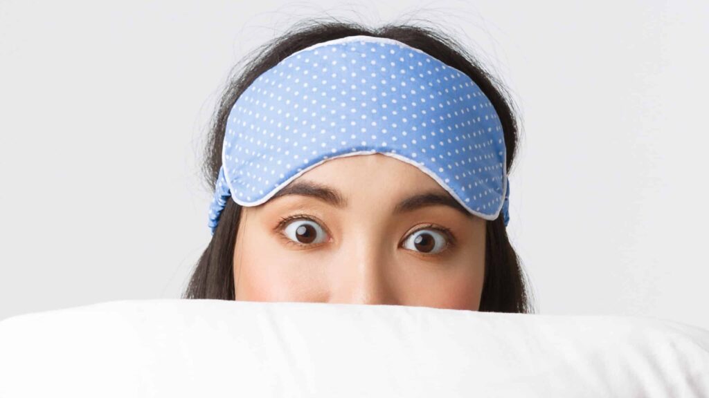 What is Rapid Eye Movement Sleep Behavior Disorder? Closeup of woman with a sleeping mask peeking over a pillow. Blog by Whitney Sleep Center